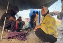 Geçici bir çadırın altında, yerde ateş üzerinde yemek hazırlanırken bir kadın gülümseyerek oturuyor, yanında bir adam küçük bir çocuğu kucağında tutuyor ve diğerleri günlük işlerle meşgul oluyor.