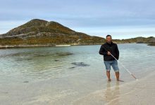 Görme engelli öğretim üyesi Dr. Önder İşlek, sahilde, berrak sığ suların içinde çıplak ayakla dururken, elindeki beyaz bastonla poz veriyor.