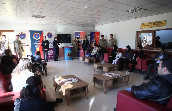 Bir salonda jandarma personelinin katılımıyla düzenlenen toplantıda, koltuklarda oturan engelli bireyler ve ailelerine yönelik bilgilendirme yapılıyor. Arka planda jandarmaya ait afişler yer alırken, ortam resmi ama sıcak bir buluşma havası taşıyor.