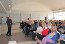 Geniş bir salonda ayakta sunum yapan bir konuşmacı, karşısında sandalyelere oturmuş çoğunluğu yaşlı bir dinleyici grubuna hitap ediyor.