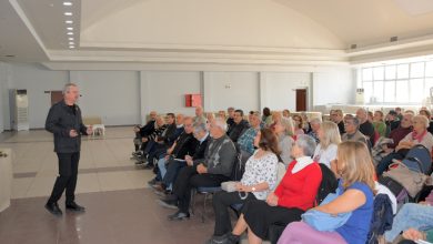 Geniş bir salonda ayakta sunum yapan bir konuşmacı, karşısında sandalyelere oturmuş çoğunluğu yaşlı bir dinleyici grubuna hitap ediyor.