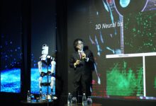 Sahnede takım elbiseli bir konuşmacı, mikrofonla sunum yapıyor. Yanında insansı bir robot ayakta duruyor. Arkadaki dev ekranda “3D Neural tissue” yazılı bilimsel görseller var.