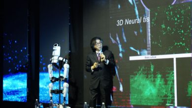 Sahnede takım elbiseli bir konuşmacı, mikrofonla sunum yapıyor. Yanında insansı bir robot ayakta duruyor. Arkadaki dev ekranda “3D Neural tissue” yazılı bilimsel görseller var.