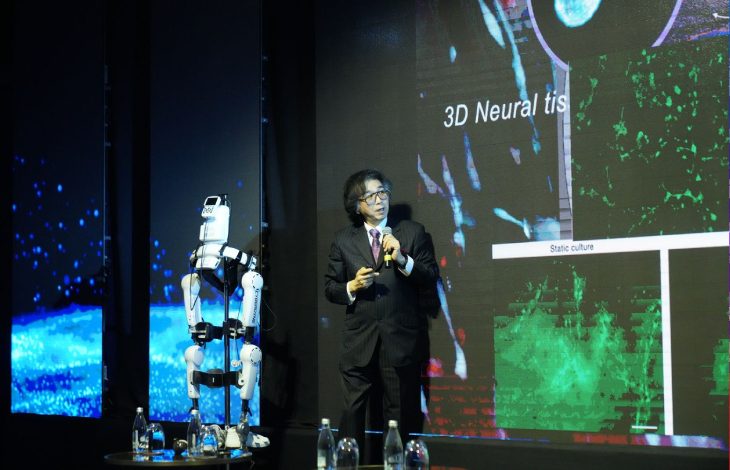 Sahnede takım elbiseli bir konuşmacı, mikrofonla sunum yapıyor. Yanında insansı bir robot ayakta duruyor. Arkadaki dev ekranda “3D Neural tissue” yazılı bilimsel görseller var.