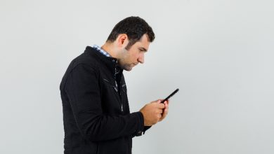 Beyaz bir duvar önünde duran bir adam, başını eğmiş şekilde telefonuna odaklanmış. Ellerinde telefonu sıkıca tutuyor, tüm dikkati ekrana kilitlenmiş. Sahne, dijital dünyaya dalıp çevreyle bağın koptuğu bir anı yakalıyor.