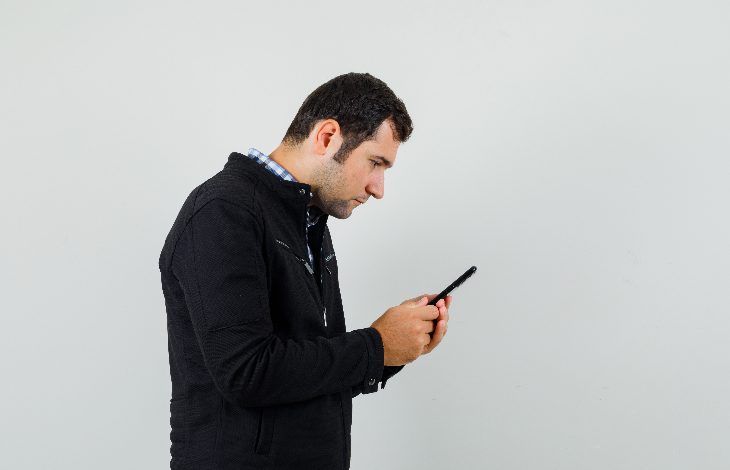 Beyaz bir duvar önünde duran bir adam, başını eğmiş şekilde telefonuna odaklanmış. Ellerinde telefonu sıkıca tutuyor, tüm dikkati ekrana kilitlenmiş. Sahne, dijital dünyaya dalıp çevreyle bağın koptuğu bir anı yakalıyor.