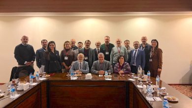 Toplantı salonunda, uzun bir masa etrafında toplanmış kalabalık bir grup görülüyor. Ön sırada oturanlar, arkada ayakta duranlarla birlikte kameraya poz vermiş. Masa üzerinde su şişeleri ve ikramlar var; ortam resmi ve kurumsal bir toplantı havasında.