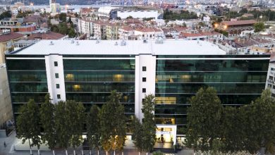 Şehrin ortasında yükselen geniş cam cepheli modern bir hastane binası; önünde sıralı ağaçlar, arka planda da dağlara yaslanmış kalabalık şehir manzarası uzanıyor.