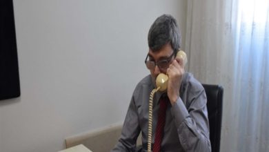 Bir ofiste masasında oturan, gözlük takan görme engelli bir erkek, sabit hatlı telefonu kulağına tutarak görüşme yapıyor; sade ve çalışma odaklı bir ortam görülüyor.