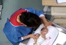 Üstten çekilmiş görüntüde, hastane ortamında bir sağlık çalışanının yenidoğan bir bebeğe bakım uyguladığı görülüyor; bebek kuvöz benzeri bir yatakta, çalışanın elleri bebeğin baş ve kol bölgesinde.