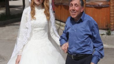 Gelinlik içindeki genç bir kadın, koluna girmiş görme engelli bir erkekle yürürken görüntüleniyor. Güneşli bir sokak, ahşap bir yapı arka planda.