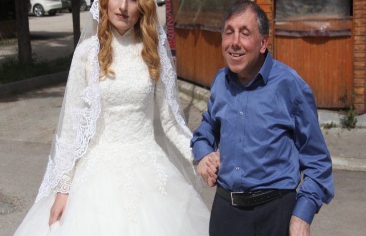 Gelinlik içindeki genç bir kadın, koluna girmiş görme engelli bir erkekle yürürken görüntüleniyor. Güneşli bir sokak, ahşap bir yapı arka planda.