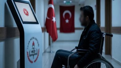 Bir kurum koridorunda, tekerlekli sandalyedeki bir kişi MEB logosu bulunan dijital bir ekrana doğru beklerken görülüyor; arka planda Türk bayrakları ve resmi, soğuk bir atmosfer hâkim.