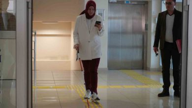 Beyaz baston kullanan bir kadın, kapalı bir mekânda sarı kılavuz çizgiler üzerinde telefonuna bakarak yürürken yanında bulunan yönetici erkek onu izliyor.