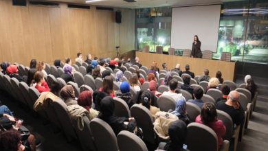 Yazar Espina Hande Ayas, kürsüde ayakta durarak konuşma yapıyor, katılımcılar salon koltuklarında oturarak konuşmayı dinliyor.