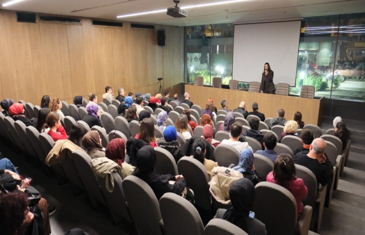 Yazar Espina Hande Ayas, kürsüde ayakta durarak konuşma yapıyor, katılımcılar salon koltuklarında oturarak konuşmayı dinliyor.