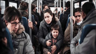 Kalabalık bir otobüste, ayakta duran bir anne kucağındaki ağlayan çocuğu sıkıca tutuyor. Etraflarındaki yolcular yorgun ve kayıtsız; dar alanda sıkışmışlık ve gerginlik hissi öne çıkıyor.