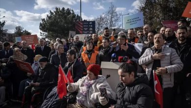 Kalabalık bir açık hava meydanında, engelli bireyler ve dernek temsilcileri bir araya gelmiş. Ön planda tekerlekli sandalyede oturan kişiler Türk bayrakları tutuyor; arkada mikrofonlu bir konuşmacı ve pankartlar yükseliyor. Yüzlerde kararlılık, havada talep ve dayanışma var.