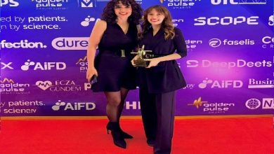 Kırmızı halıda iki kadın yan yana poz veriyor. İkisi de siyah kıyafetli; biri elinde ödül tutuyor. Arkalarında mor tonlarda, sponsor logolarıyla dolu bir ödül töreni fotoğraf duvarı var.