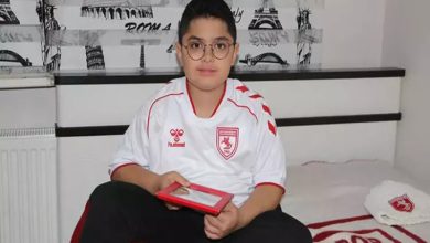 Bir odada oturan gözlüklü bir çocuk, Samsunspor formasıyla elinde tablet tutuyor; arkasında desenli duvar kâğıdı ve yatağı görülüyor.