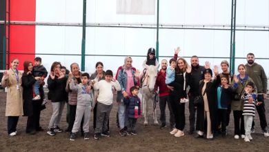 Kapalı bir alanda, farklı yaşlardan yetişkinler ve çocuklar yan yana dizilmiş, kameraya el sallıyor. Ortada bir çocuk ata binmiş; yüzlerde gülümseme, birlikte olmanın sıcak ve destekleyici havası öne çıkıyor.