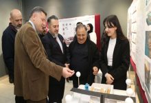 Bir sergi alanında, birkaç kişi mimari bir maket masasının etrafında toplanmış. Ön tarafta genç bir kadın, hazırladığı proje maketini anlatıyor; karşısında takım elbiseli ve ceketli erkekler dikkatle inceleyip sohbet ediyor. Arkada panolara asılmış mimari proje sunumları var.