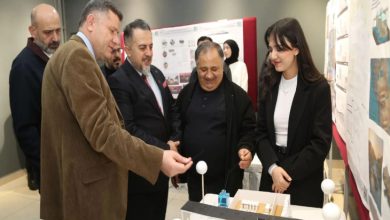 Bir sergi alanında, birkaç kişi mimari bir maket masasının etrafında toplanmış. Ön tarafta genç bir kadın, hazırladığı proje maketini anlatıyor; karşısında takım elbiseli ve ceketli erkekler dikkatle inceleyip sohbet ediyor. Arkada panolara asılmış mimari proje sunumları var.