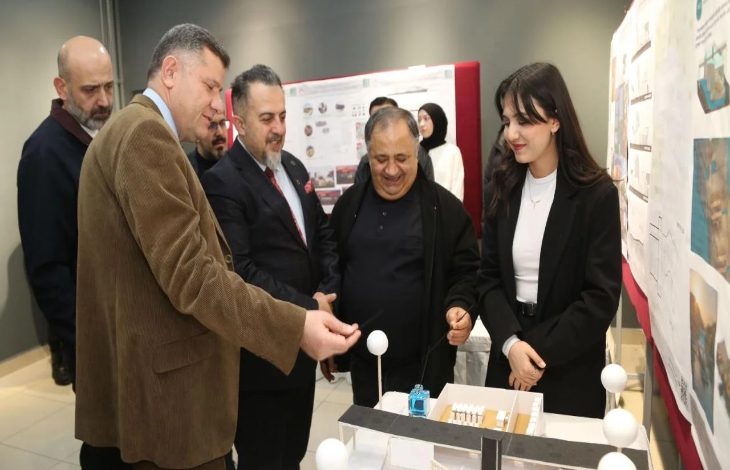 Bir sergi alanında, birkaç kişi mimari bir maket masasının etrafında toplanmış. Ön tarafta genç bir kadın, hazırladığı proje maketini anlatıyor; karşısında takım elbiseli ve ceketli erkekler dikkatle inceleyip sohbet ediyor. Arkada panolara asılmış mimari proje sunumları var.