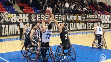 Kapalı bir spor salonunda, tekerlekli sandalyedeki basketbolcuların hava atışı için topa uzandığı, tribünleri dolu ve rekabetin yoğun olduğu bir tekerlekli sandalye basketbol maçı görülüyor.
