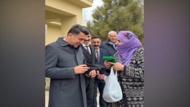 Açık havada, resmi giyimli bir adam yaşlı bir kadının uzattığı el emeği örgüyü inceliyor; etraflarında birkaç kişi gülümseyerek anı izliyor, sahne samimi ve sıcak.