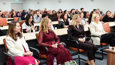 Bir konferans salonunda, ön sıralarda oturan katılımcılar dikkatle bir sunumu dinliyor. Arkada sıralar dolu, ortam resmi ve bilgilendirici bir toplantı havasında.
