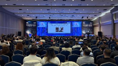 Büyük bir kongre salonunda, sıralar halinde oturmuş kalabalık bir katılımcı grubu görülüyor. Sahnedeki dev ekranda sunum yansıtılıyor; yan ekranlarda konuşmacının görüntüsü yer alıyor. Ortam profesyonel, akademik bir kongre havasında.