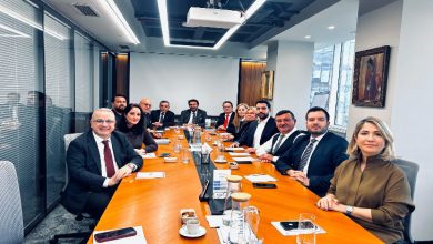 Uzun bir toplantı masası etrafında dizilmiş resmi giyimli bir grup insan kameraya dönük şekilde poz veriyor. Masada notlar, telefonlar ve kahveler; arka planda modern bir ofis ve şehir manzarası.