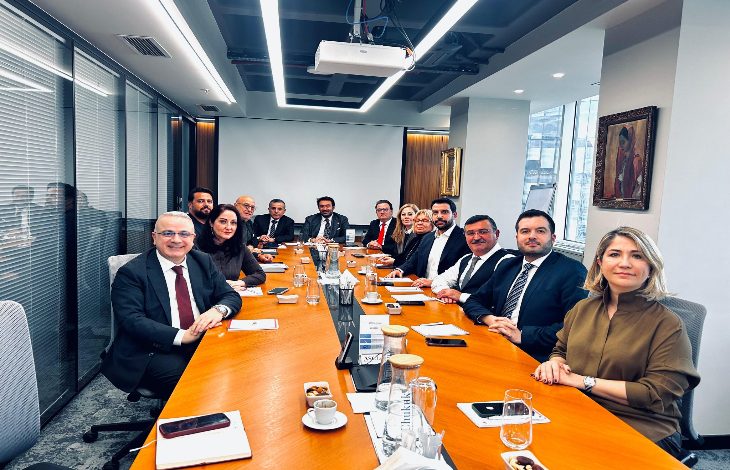 Uzun bir toplantı masası etrafında dizilmiş resmi giyimli bir grup insan kameraya dönük şekilde poz veriyor. Masada notlar, telefonlar ve kahveler; arka planda modern bir ofis ve şehir manzarası.