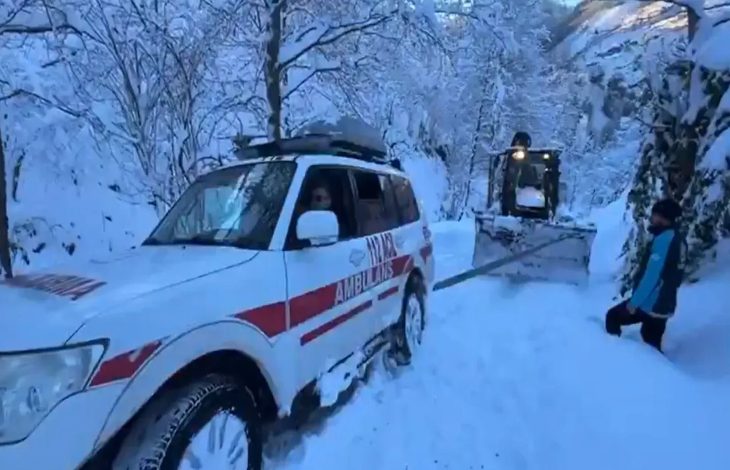 Yoğun karla kaplı bir yolda, kar küreme aracının açtığı güzergahta ilerleyen bir ambulans görülüyor; çevre bembeyaz, ekipler zorlu kış koşullarında müdahale halinde.