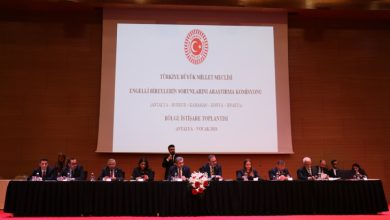 Büyük bir konferans salonunda, Türkiye Büyük Millet Meclisi Engelli Bireylerin Sorunlarını Araştırma Komisyonu’nun Antalya’da düzenlenen bölge istişare toplantısında, uzun bir masada oturan katılımcılar ve arkadaki resmi sunum ekranı görülüyor.