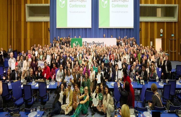 Büyük bir konferans salonunda yüzlerce kişinin yer aldığı toplu bir hatıra fotoğrafı çekilmiş. Arkada “Zero Project Conference” yazılı büyük ekranlar bulunuyor. Katılımcılar sahne önünde ve salon içinde bir araya gelmiş; bazıları sertifika ya da plaket tutuyor. Uluslararası ve kapsayıcı bir etkinliğin coşkulu final karesi gibi görünüyor.