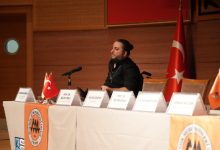 Bir konferans salonunda, önünde mikrofon bulunan bir masada oturan bir konuşmacı görülüyor. Masanın üzerinde isimlikler ve küçük Türk bayrakları yer alırken, arka planda büyük bir Türk bayrağı asılı; ortam resmi bir panel ya da toplantı havası taşıyor.