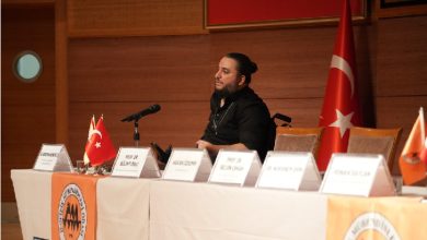 Bir konferans salonunda, önünde mikrofon bulunan bir masada oturan bir konuşmacı görülüyor. Masanın üzerinde isimlikler ve küçük Türk bayrakları yer alırken, arka planda büyük bir Türk bayrağı asılı; ortam resmi bir panel ya da toplantı havası taşıyor.