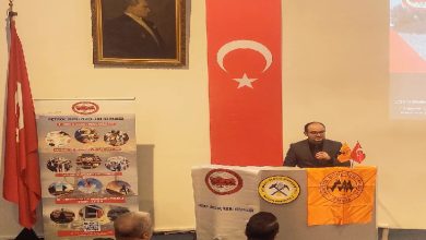 Bir konferans salonunda kürsüde konuşma yapan bir erkek görülüyor. Kürsünün önünde çeşitli dernek logolarının yer aldığı flamalar asılı. Arka duvarda Türk bayrağı ve Mustafa Kemal Atatürk’ün portresi bulunuyor. Sol tarafta bir derneğe ait tanıtım afişi yer alırken, dinleyiciler ön sırada oturmuş şekilde konuşmayı takip ediyor. Ortam resmi bir panel ya da anma programı izlenimi veriyor.