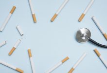 Açık mavi zemin üzerine dağınık şekilde yerleştirilmiş çok sayıda sigara görülüyor. Sağ tarafta bir stetoskop bulunuyor. Görsel, sigara kullanımının sağlık üzerindeki etkilerine ve tıbbi boyutuna dikkat çeken sembolik bir kompozisyon sunuyor.