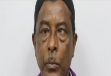 Yakın plan çekilmiş bir erkek portresi görülüyor. Kişi doğrudan kameraya bakıyor. Koyu renk bir bere takıyor ve mor tonlarında bir üst giysi giymiş. Yüz ifadesi ciddi ve nötr; arka plan ise düz, açık renkli ve sade.