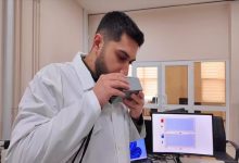 Bir laboratuvar ortamında beyaz önlüklü bir araştırmacı, elindeki küçük cihazı ağzına yaklaştırarak nefes üflüyor. Yan tarafta bilgisayar ekranında ölçüm sonuçlarını gösteren grafikler yer alıyor. Ortam akademik bir çalışma ve test sürecini yansıtıyor.