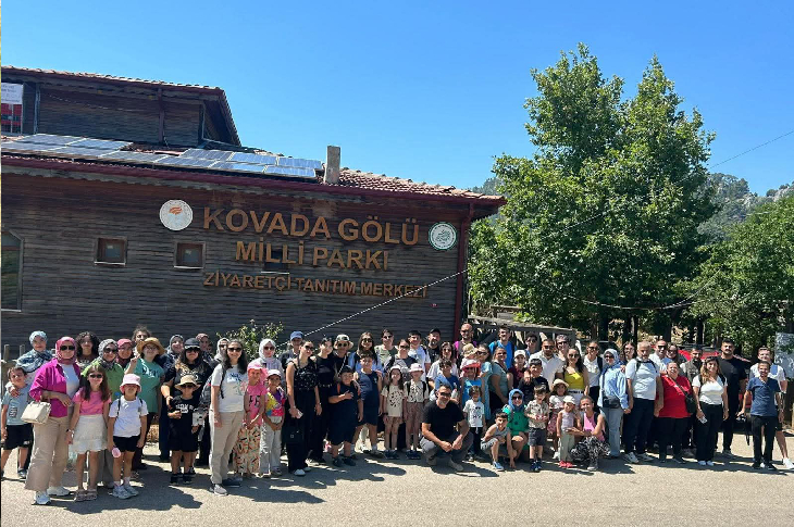 Ahşap bir bina önünde kalabalık bir grup toplu fotoğraf çektiriyor. Binanın üzerinde “Kovada Gölü Milli Parkı Ziyaretçi Tanıtım Merkezi” yazısı yer alıyor. Çoğunluğu ailelerden oluşan grupta çok sayıda çocuk da var. Hava güneşli; arka planda ağaçlar ve mavi gökyüzü görülüyor. Atmosfer bir gezi ya da toplu etkinlik havası taşıyor.