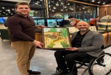 Bir pastane/kafe ortamında iki erkek yan yana poz veriyor. Ayakta duran kişi, tekerlekli sandalyede oturan kişiye çerçeveli bir tablo takdim ediyor. Tabloda taş evler ve çiçekli bir sokak resmi yer alıyor; arka planda tatlı tezgâhları ve çalışanlar görülüyor.