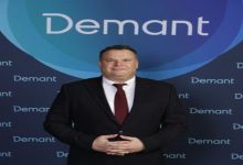 Koyu renk takım elbise ve bordo kravat takan bir erkek, üzerinde “Demant” logosu bulunan mavi tonlardaki bir fon önünde ayakta duruyor. Ellerini önde birleştirmiş, kameraya doğru sakin ve resmi bir poz veriyor.