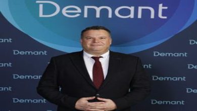 Koyu renk takım elbise ve bordo kravat takan bir erkek, üzerinde “Demant” logosu bulunan mavi tonlardaki bir fon önünde ayakta duruyor. Ellerini önde birleştirmiş, kameraya doğru sakin ve resmi bir poz veriyor.