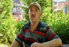 Açık havada, yeşillikler arasında oturan yaşlı bir adam görülüyor. Üzerinde çizgili tişört ve şapka var; sakin ama yorgun bir ifadeyle kameraya bakıyor. Arka planda ağaçlar ve şehir yapıları seçiliyor.