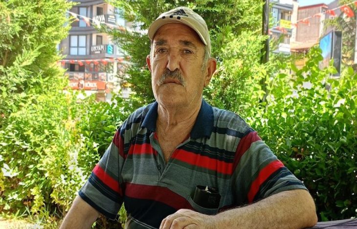 Açık havada, yeşillikler arasında oturan yaşlı bir adam görülüyor. Üzerinde çizgili tişört ve şapka var; sakin ama yorgun bir ifadeyle kameraya bakıyor. Arka planda ağaçlar ve şehir yapıları seçiliyor.