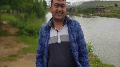 Bir nehir kıyısında açık havada çekilmiş bir fotoğraf. Önde duran bir erkek kameraya gülümseyerek poz veriyor. Üzerinde mavi mont ve polo yaka bir tişört var. Arka planda su kenarı, yeşillik alan ve bir köprü görünüyor.